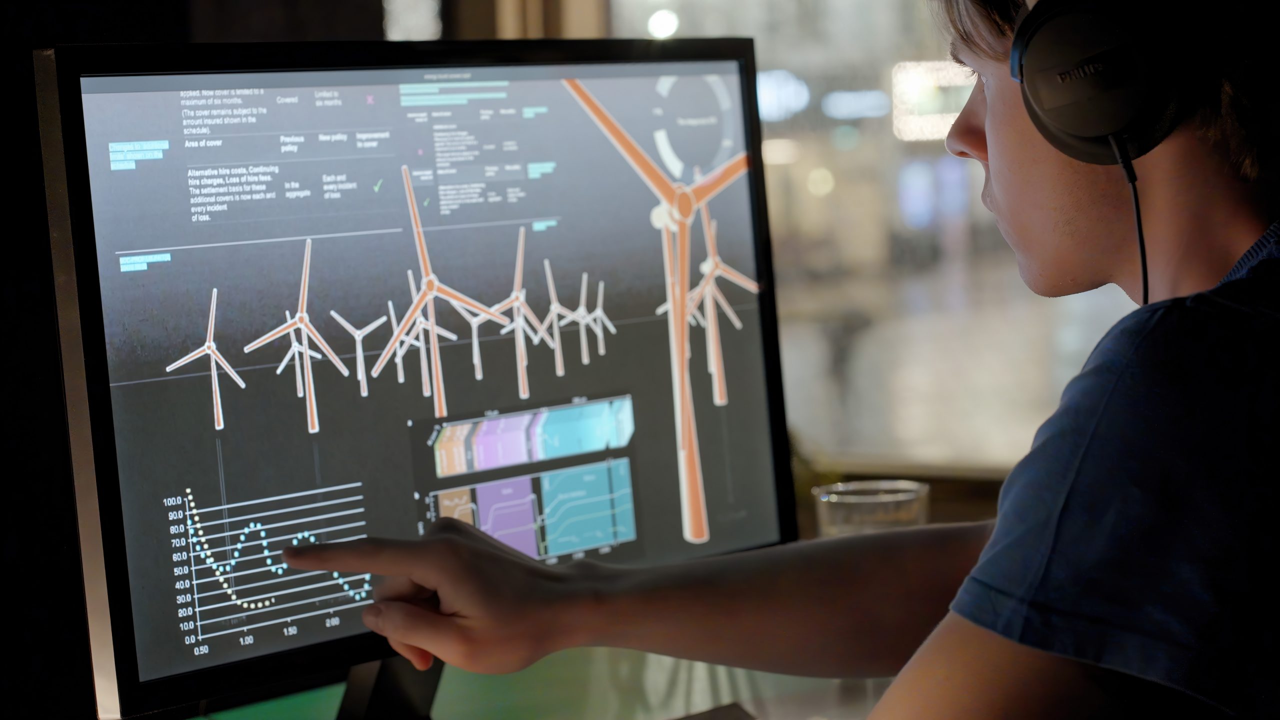young man, possibly a student, working with a touchscreen computer at night. He’s researching data relating to sustainable energy production, the background is a rain drenched urban scene.